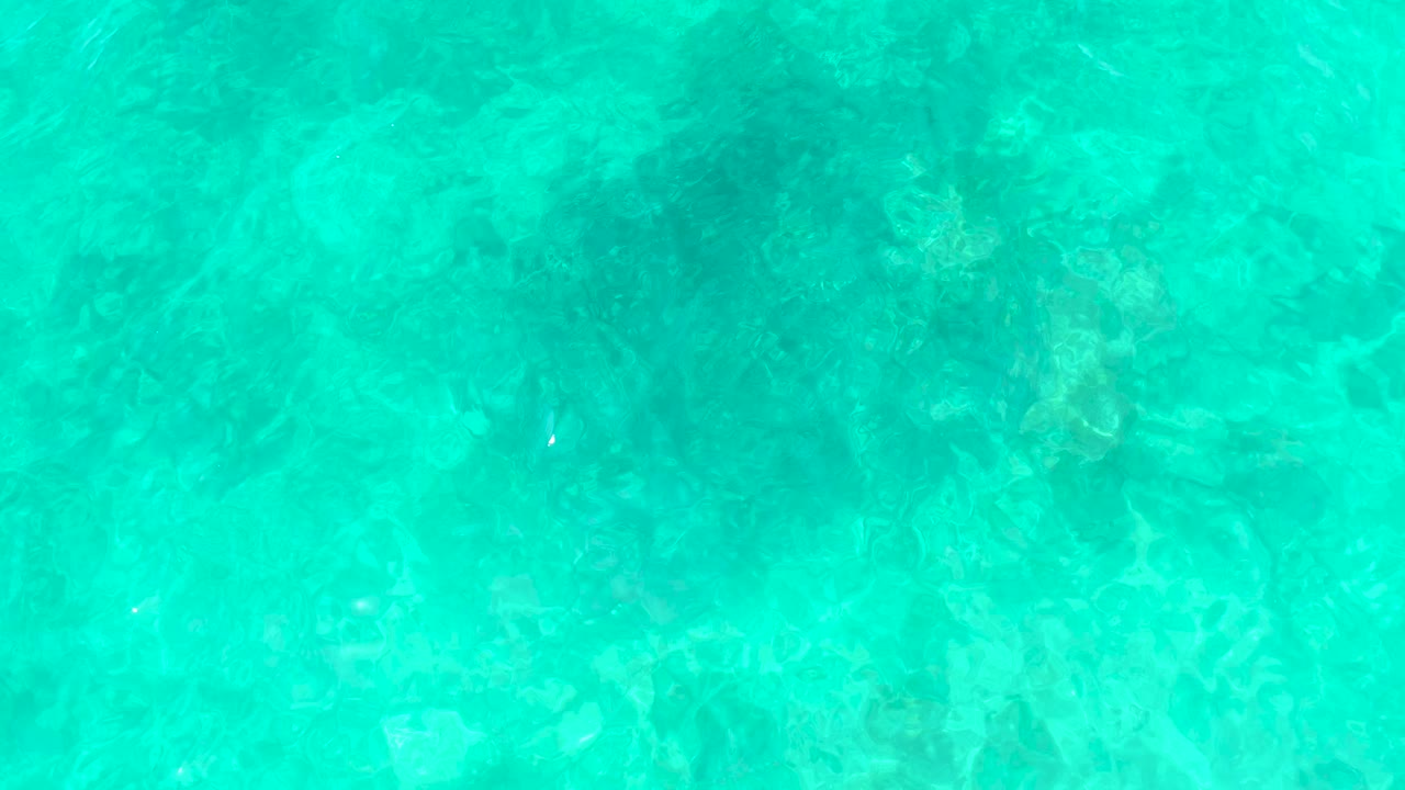 Top-down view of clear turquoise water at El Tule Beach in Los Cabos with faint seabed texture below and no visible people.