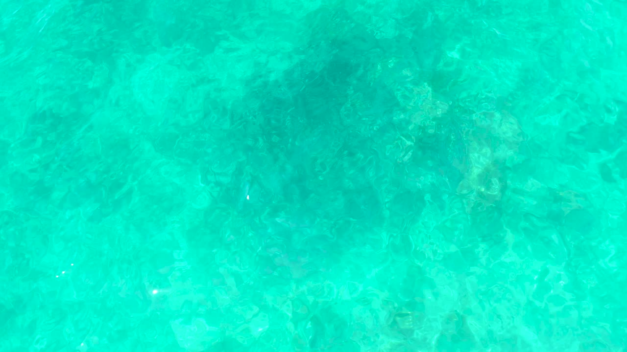 Clear turquoise ocean surface at El Tule Beach in Los Cabos shows darker coral or rock patches beneath small ripples, with no visible people.