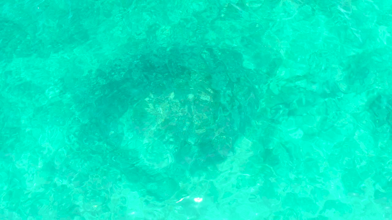Overhead view of clear turquoise water at El Tule Beach in Los Cabos with darker submerged rocks beneath the rippled surface and no visible people.