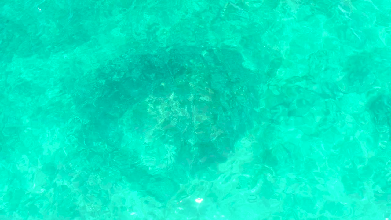 Clear blue-green water fills the frame at El Tule Beach in Los Cabos, with mottled underwater rock shapes softened by ripples and no visible people.