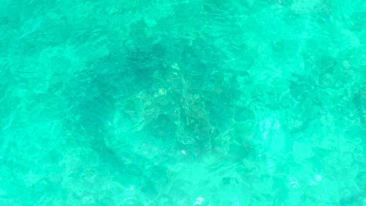 Bright turquoise water at El Tule Beach in Los Cabos reveals faint underwater formations and scattered light glints across the rippled surface, with no visible people.