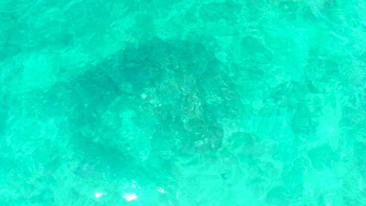 Transparent turquoise ocean at El Tule Beach in Los Cabos shows darker rock clusters under the moving surface texture and no visible people.