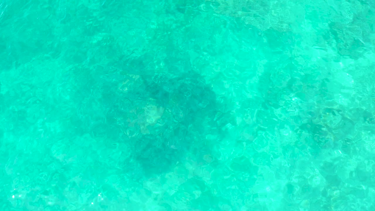 Bright overhead view of El Tule Beach Los Cabos water with the rocky bottom visible through ripples and no visible people.