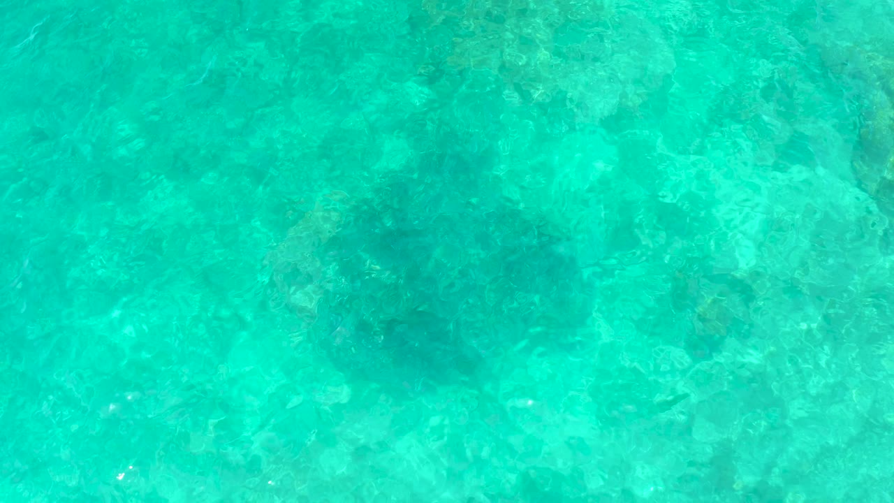 Clear turquoise ocean water at El Tule Beach Los Cabos shows a broad dark submerged shape under soft surface texture with no visible people.