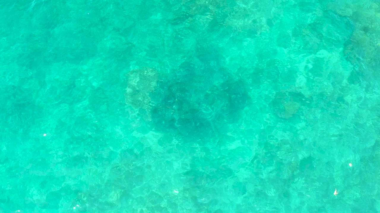 At 0:01, the El Tule Beach water remains a top-down turquoise field with mottled darker underwater patches and small bright surface reflections, with no visible people.