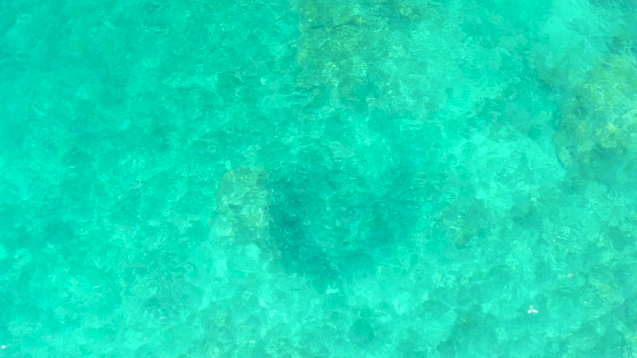 Blue-green shallow water shows pale rock shapes and darker underwater patches at El Tule Beach Los Cabos, with no visible people.