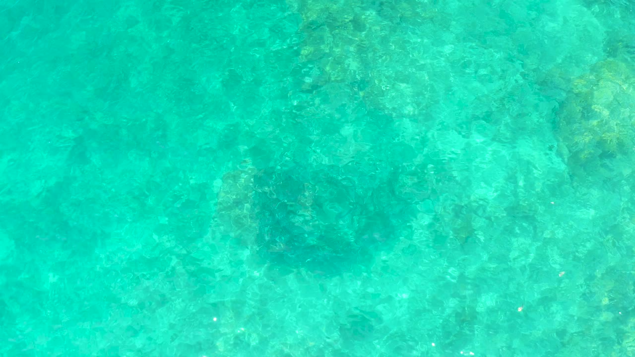 Transparent turquoise ocean water reveals clustered darker seabed areas and lighter rock below at El Tule Beach Los Cabos, with no visible people.