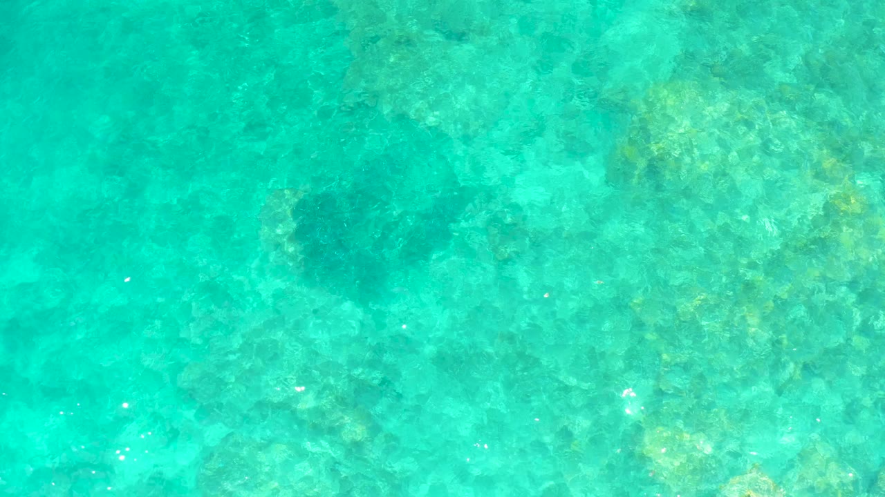 A top-down aerial frame from El Tule Beach Los Cabos shows transparent turquoise water over coral-like and rocky seabed texture, with no visible people.
