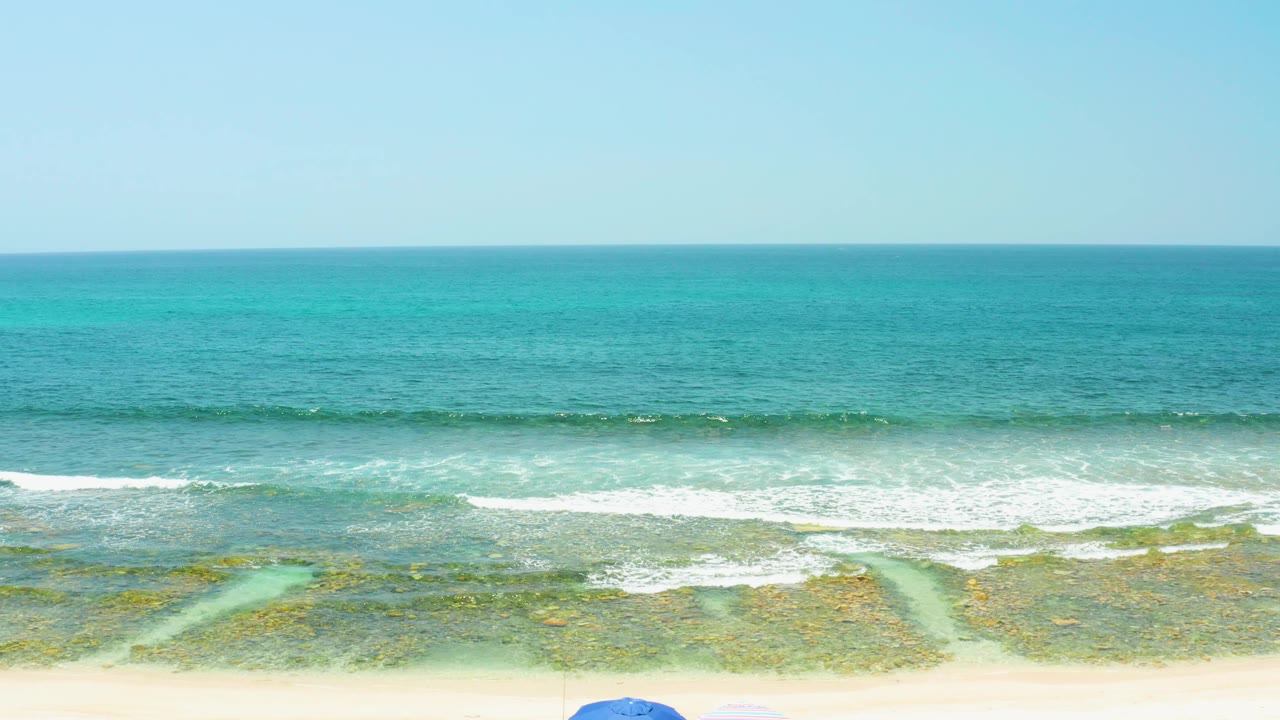 Wide elevated view of El Tule Beach Los Cabos with turquoise ocean, white surf, pale sand, and only the tops of beach umbrellas visible at the bottom edge.