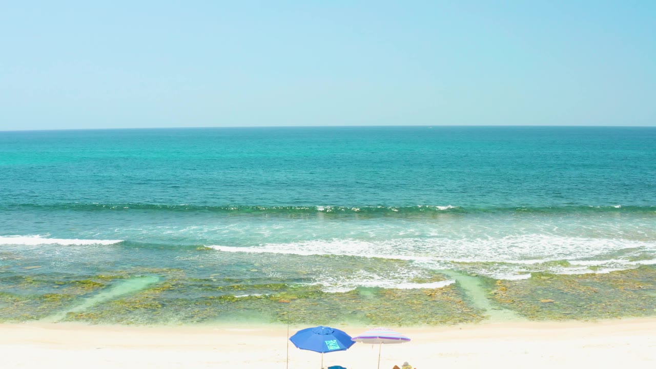 Elevated view of El Tule Beach Los Cabos showing turquoise water, white surf bands, sandy beach, and blue and striped umbrellas with beach items below.