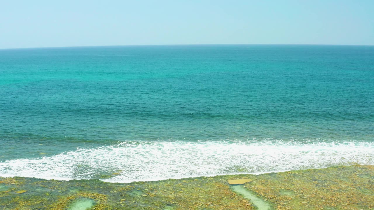 At 0:02, a darker swell band approaches the white breaking surf at El Tule Beach Los Cabos, with algae-toned rocks visible along the bottom and no visible people.