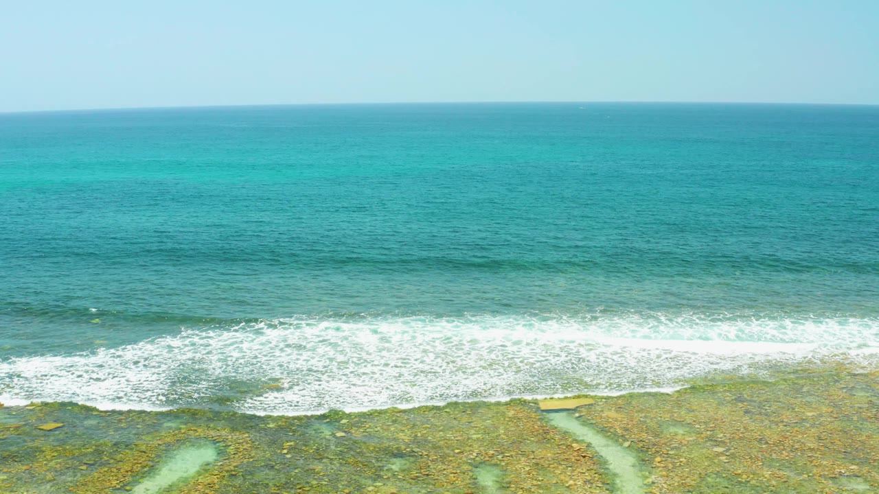 At 0:03, white water rolls unevenly along the rocky El Tule Beach Los Cabos shoreline, with clear blue-green ocean filling most of the frame and no visible people.