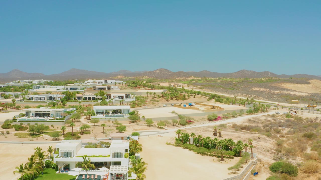 Opening frame over El Tule Beach Los Cabos shows white modern homes in sandy desert terrain with palms, distant mountains, and no visible people.