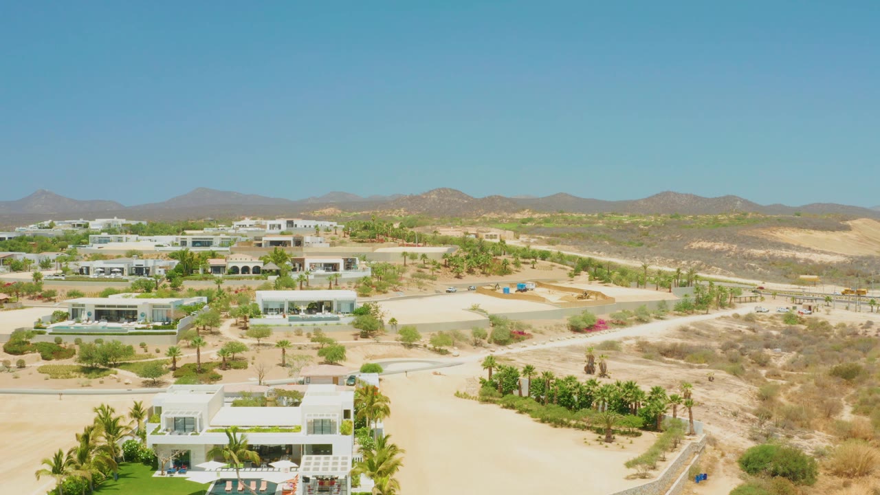 At 0:01, the aerial view in El Tule Beach Los Cabos holds on white buildings, crossing sandy roads, green plantings, dry hills, and no visible people.