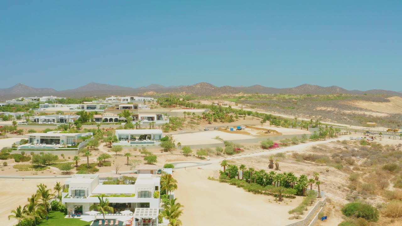 At 0:02, El Tule Beach Los Cabos appears as a sunlit desert development with pale roads, modern white homes, palms, mountains, and no visible people.