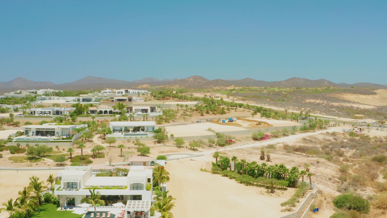 Aerial view across white houses, palm-lined landscaping, sandy lots, and mountains.