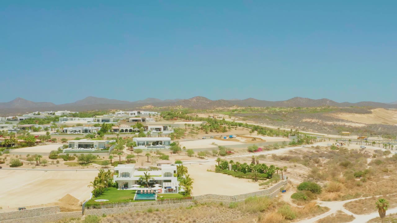 Opening aerial frame over El Tule Beach, Los Cabos, showing white modern houses, green palms, blue pools, sandy terrain, distant mountains, and no visible people.