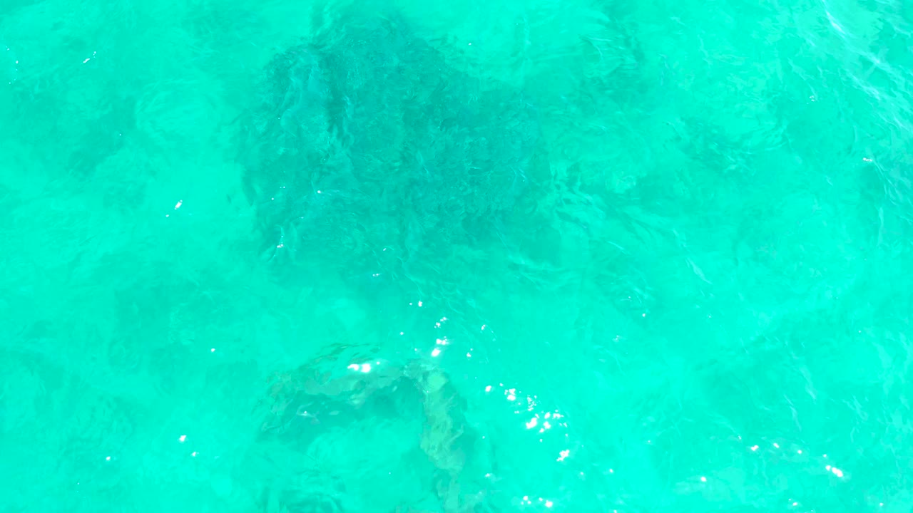 Clear aqua water at El Tule Beach Los Cabos shows submerged dark rock shapes slightly left of center, small bright reflections, and no visible people.