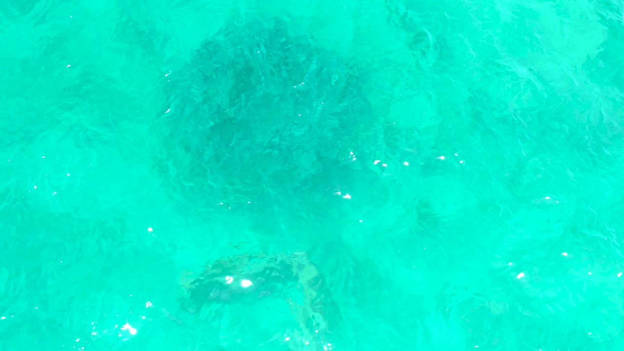 Turquoise ocean water at El Tule Beach Los Cabos reveals a darker submerged rock or coral patch near the lower right, with rippled surface light and no visible people.