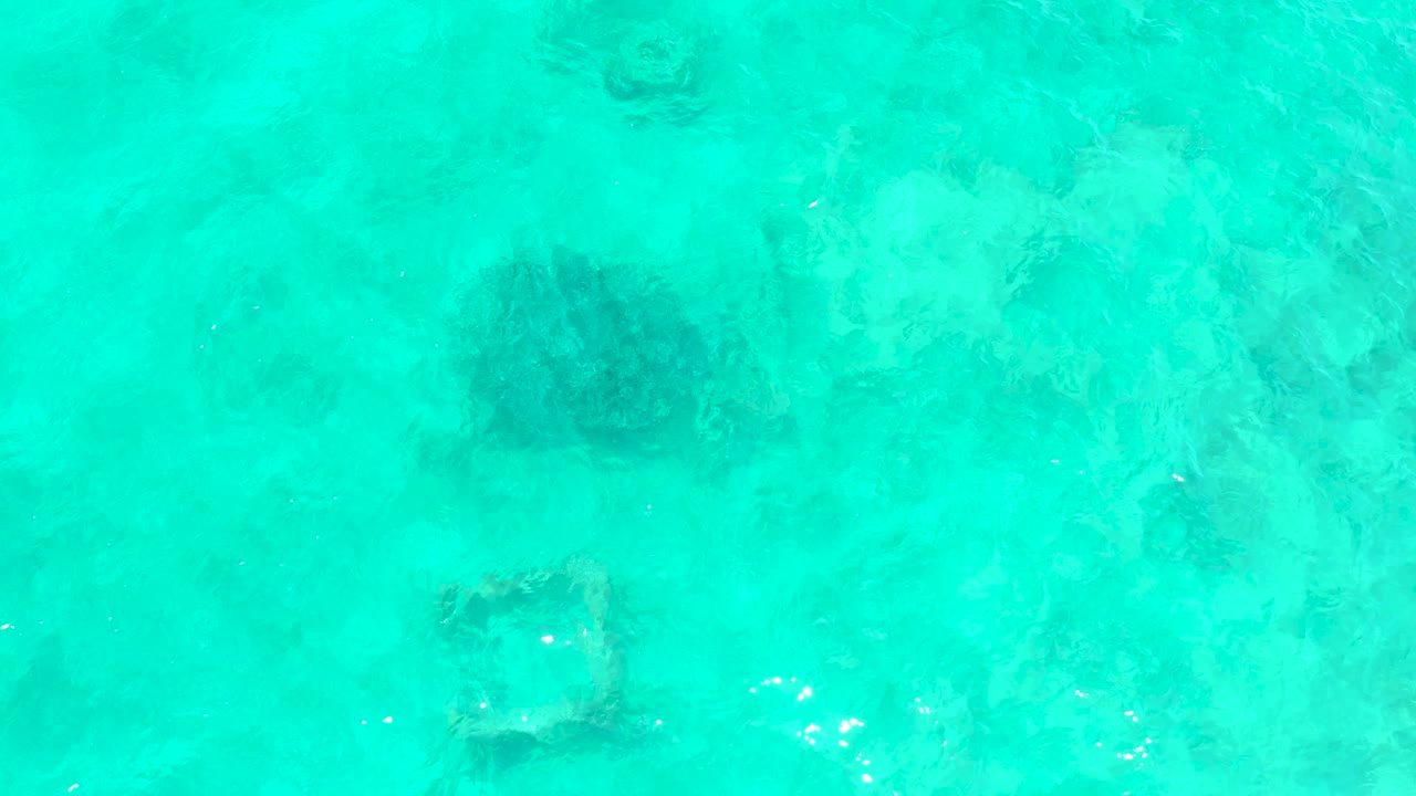 Turquoise water at El Tule Beach Los Cabos shows the same dark rounded underwater mass and square frame, with small bright ripple highlights shifting across the center and no visible people.