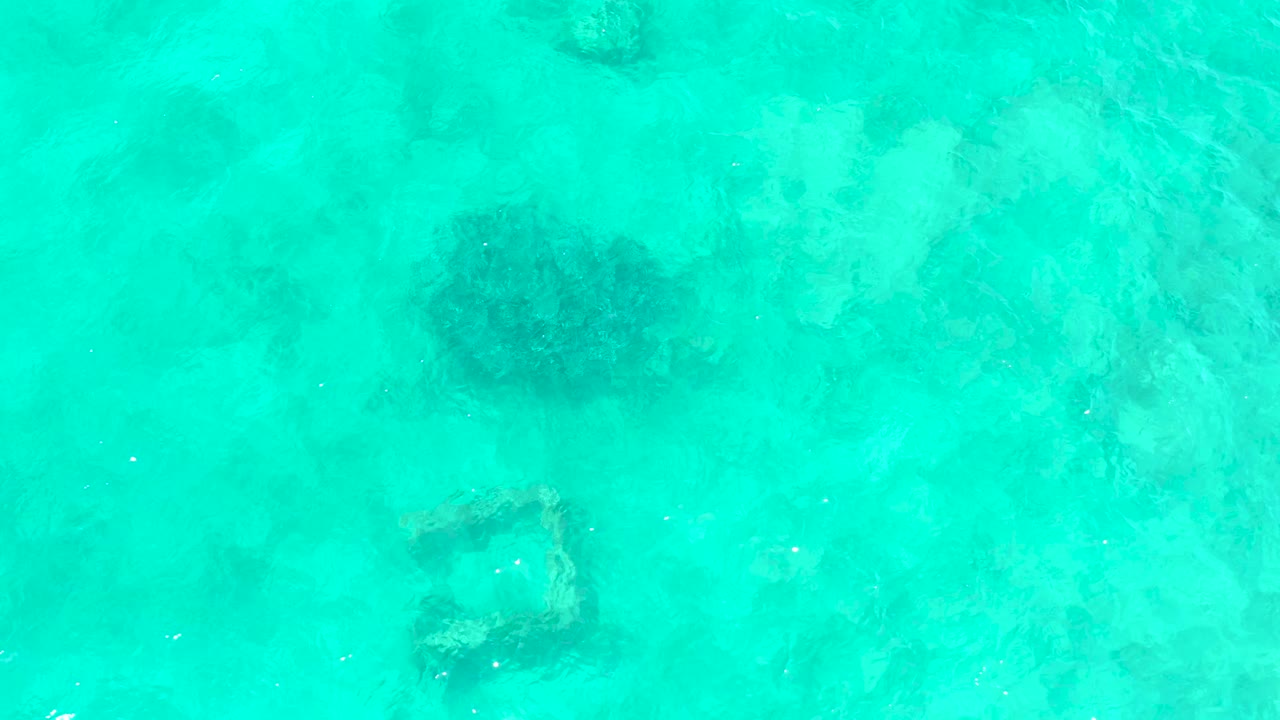 At El Tule Beach Los Cabos, the frame remains a top-down view into shallow turquoise water, where the dark rounded formation sits above a square submerged structure beneath gentle surface texture and no visible people.