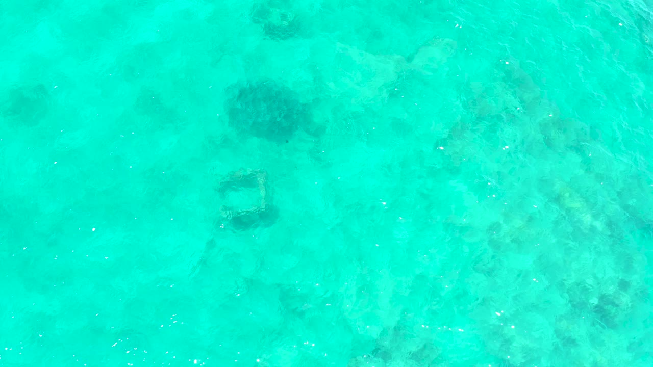 A submerged square frame shape appears beneath clear turquoise water at El Tule Beach, surrounded by pale seabed and darker marine patches with no visible people.