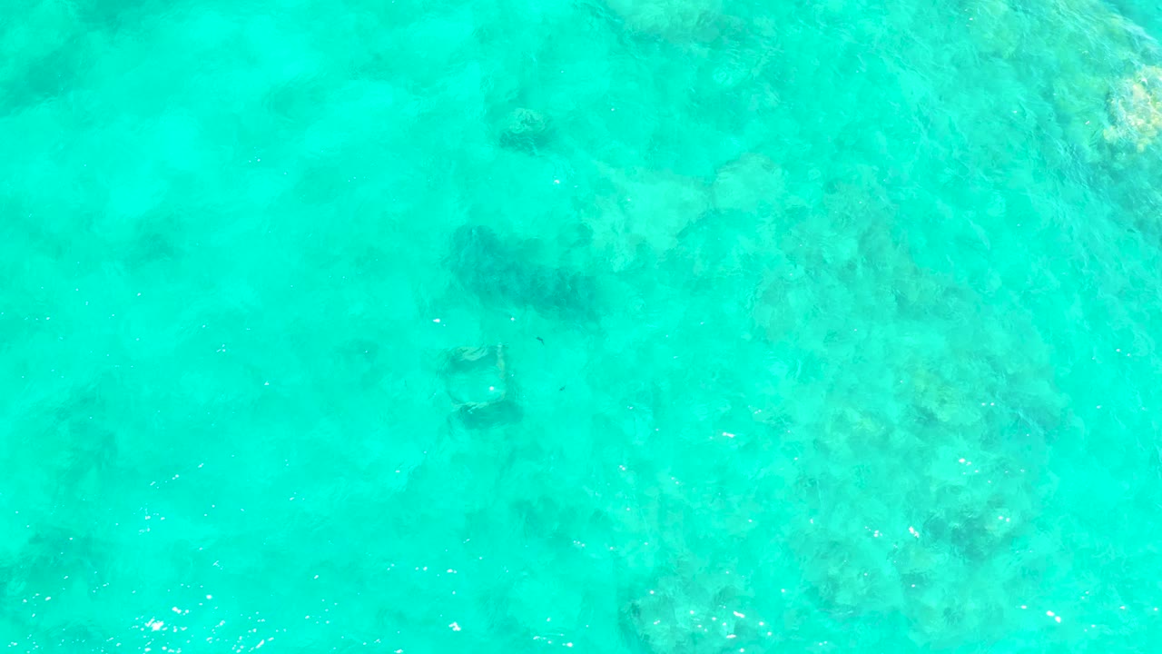 A top-down frame of shallow turquoise water at El Tule Beach, Los Cabos, with rocky patches visible through the surface and no visible people.