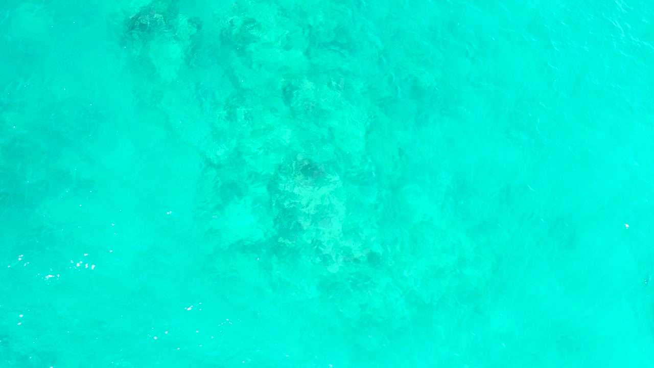 Pale turquoise ocean at El Tule Beach Los Cabos covers darker submerged rocks near the upper right, with small white surface glints and no visible people.