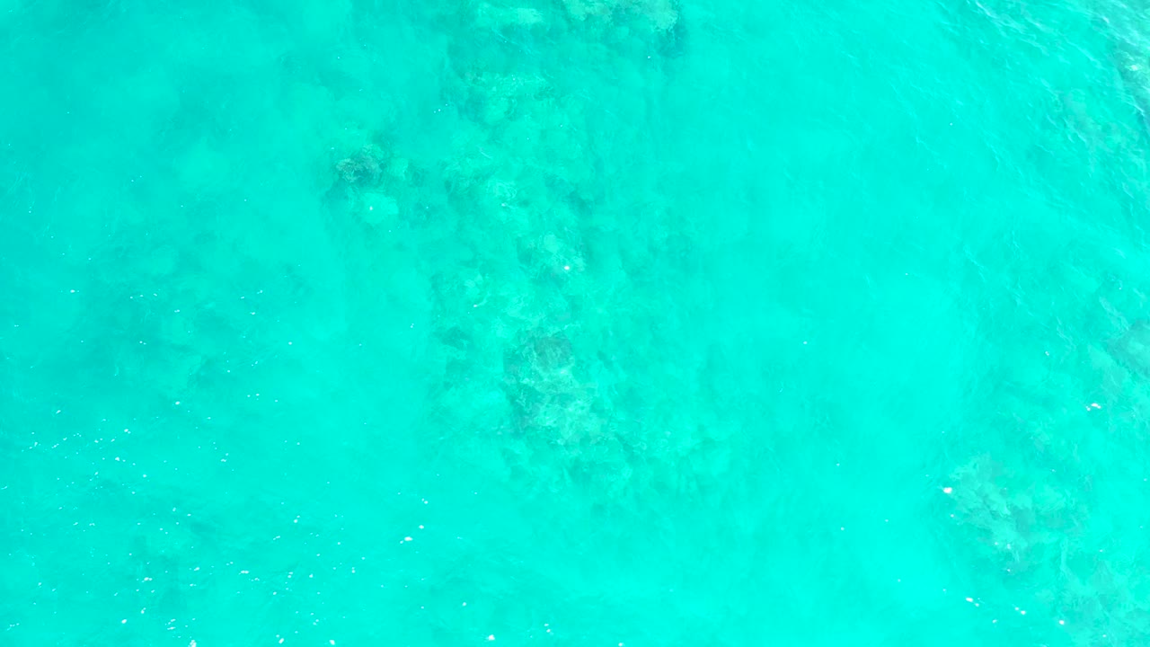 Transparent blue-green ocean water at El Tule Beach in Los Cabos reveals darker submerged patches along the right side and no visible people or objects.