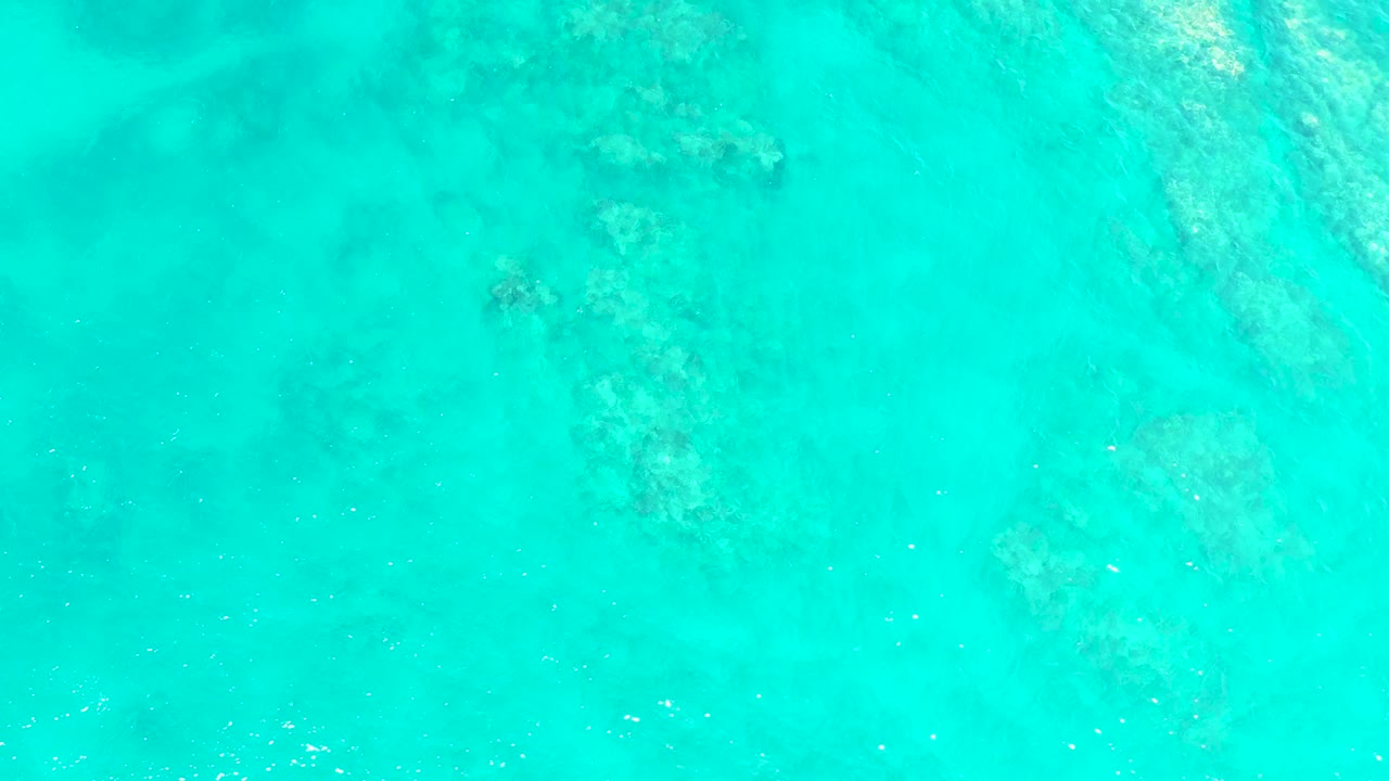 Overhead turquoise water at El Tule Beach Los Cabos contains scattered darker seabed formations and fine surface texture, with no visible people.