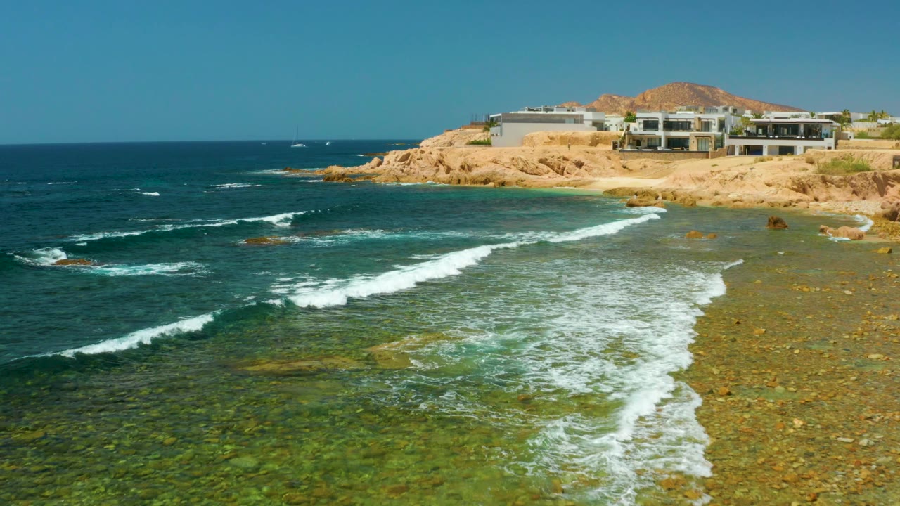 At El Tule Beach Los Cabos, clear green-blue water breaks into white foam over a rocky seabed below tan cliffs and modern buildings, with no visible people.