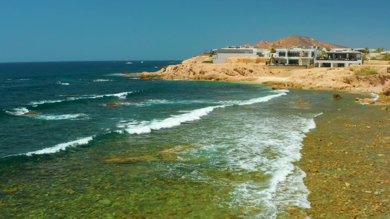 White foam spreading across shallow blue-green water near tan cliffs