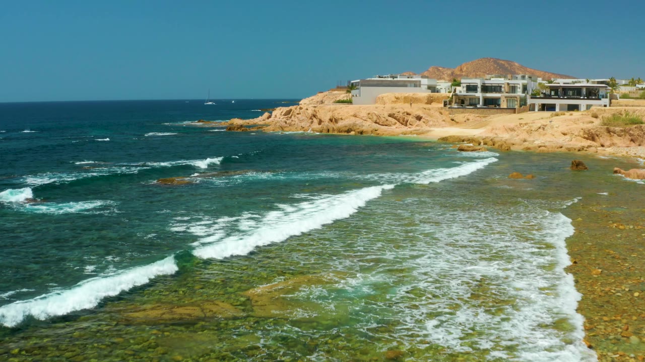 At El Tule Beach Los Cabos, foamy surf crosses the center of the clear water while the cliffside buildings sit higher on the right side of frame, with no visible people.
