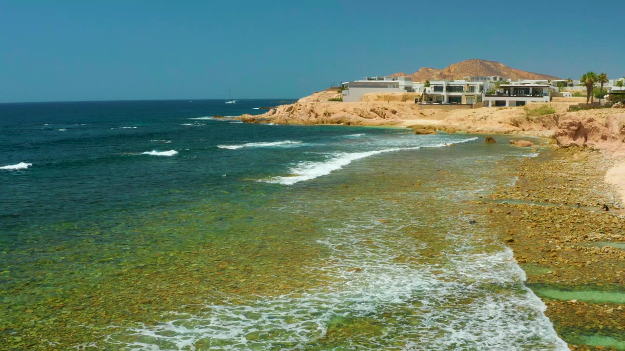 At El Tule Beach in Los Cabos, white surf spreads across shallow rocky water below tan cliffs and modern buildings, with no visible people.