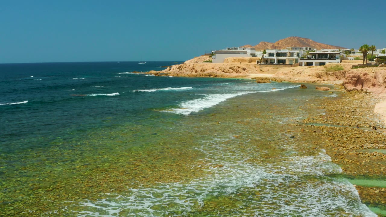 Foam breaks over green-blue shallow water at El Tule Beach, with a rocky point and buildings on the cliff in the background and no visible people.