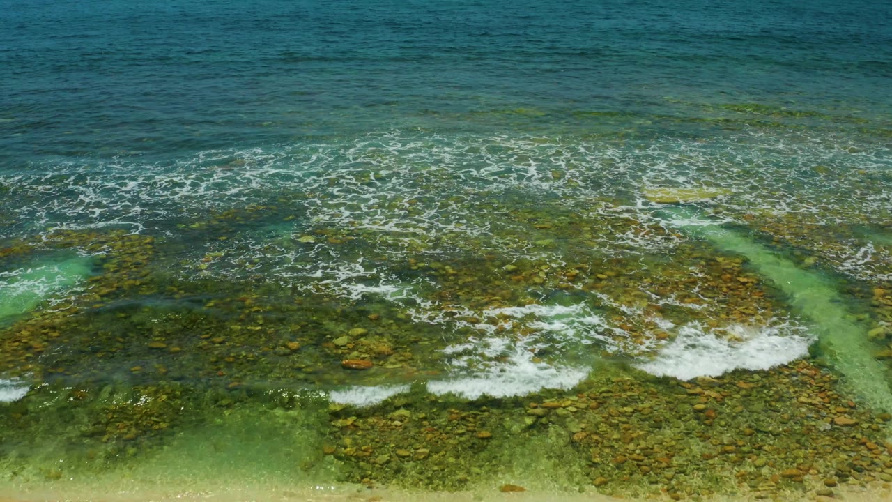 At one second, El Tule Beach shallows show a brighter patch of white foam crossing the rocky bottom beside a pale turquoise flow, with no visible people.