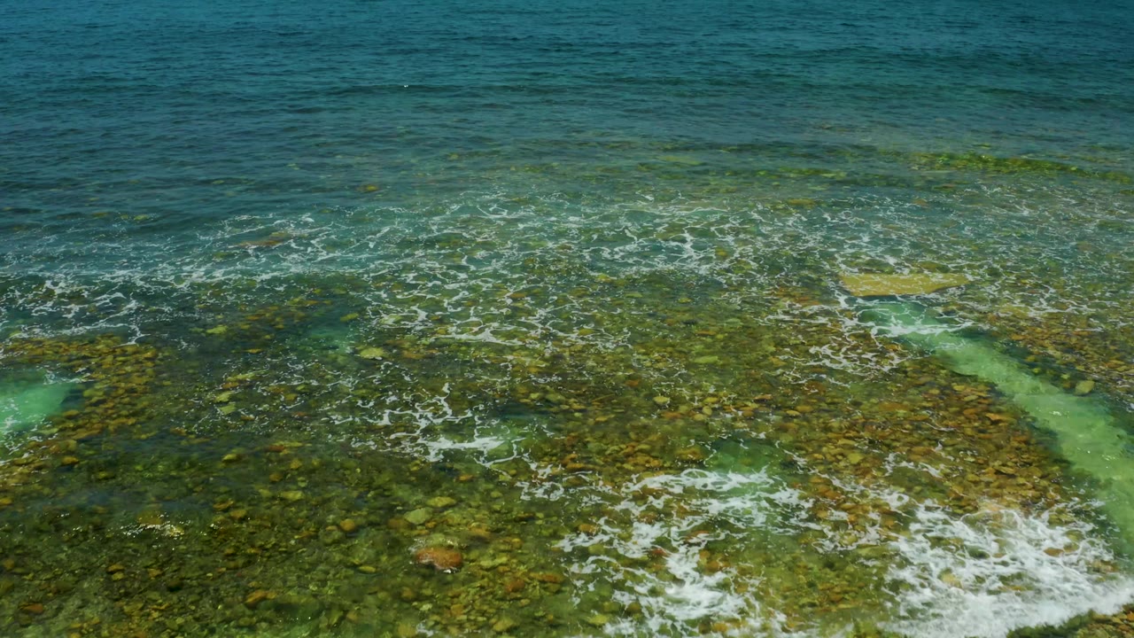 At two seconds, the aerial view holds on El Tule Beach water as surf breaks across the rocks and the clear shallows reveal the seabed, with no visible people.