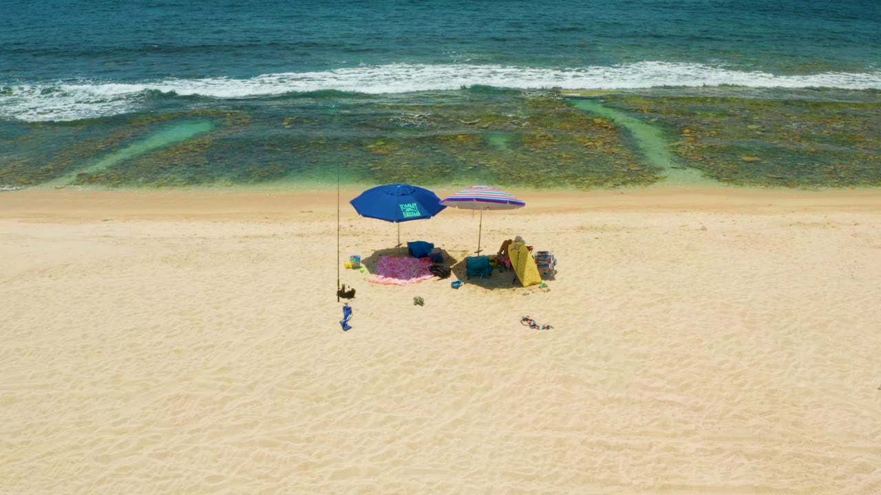 Aerial frame from El Tule Beach Los Cabos with the umbrella cluster centered below dark nearshore reef patches and a white wave line, with no visible people.