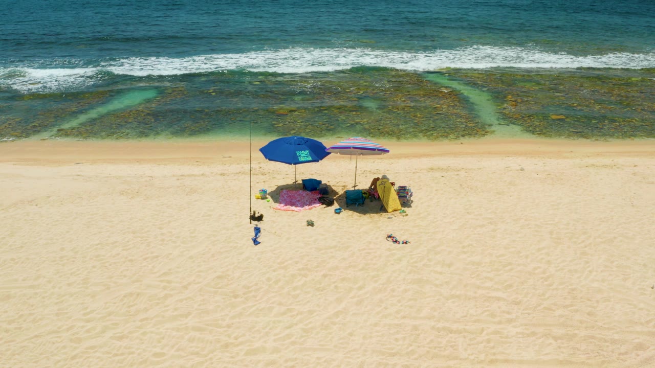 Overhead El Tule Beach Los Cabos view showing beach umbrellas on open sand, green-blue water channels through reef texture, and white surf near the top, with no visible people.