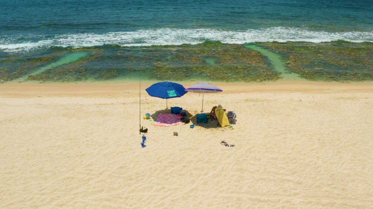 Aerial view at El Tule Beach Los Cabos with umbrellas near the lower left, broad pale sand across the frame, and foamy surf over dark reef patches, with no visible people.