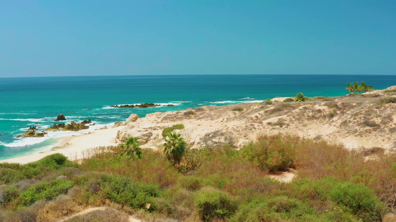 Early frame at Dreams Los Cabos showing more of the sandy path through low dune vegetation, with rock outcrops in the surf and no visible people.