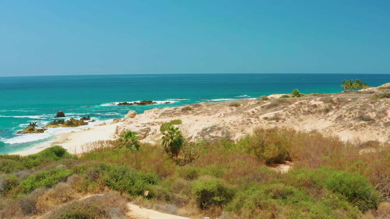 The Dreams Los Cabos beach view holds on tan dunes and green-brown scrub, with a bright ocean horizon, white surf around rocks, and no visible people.