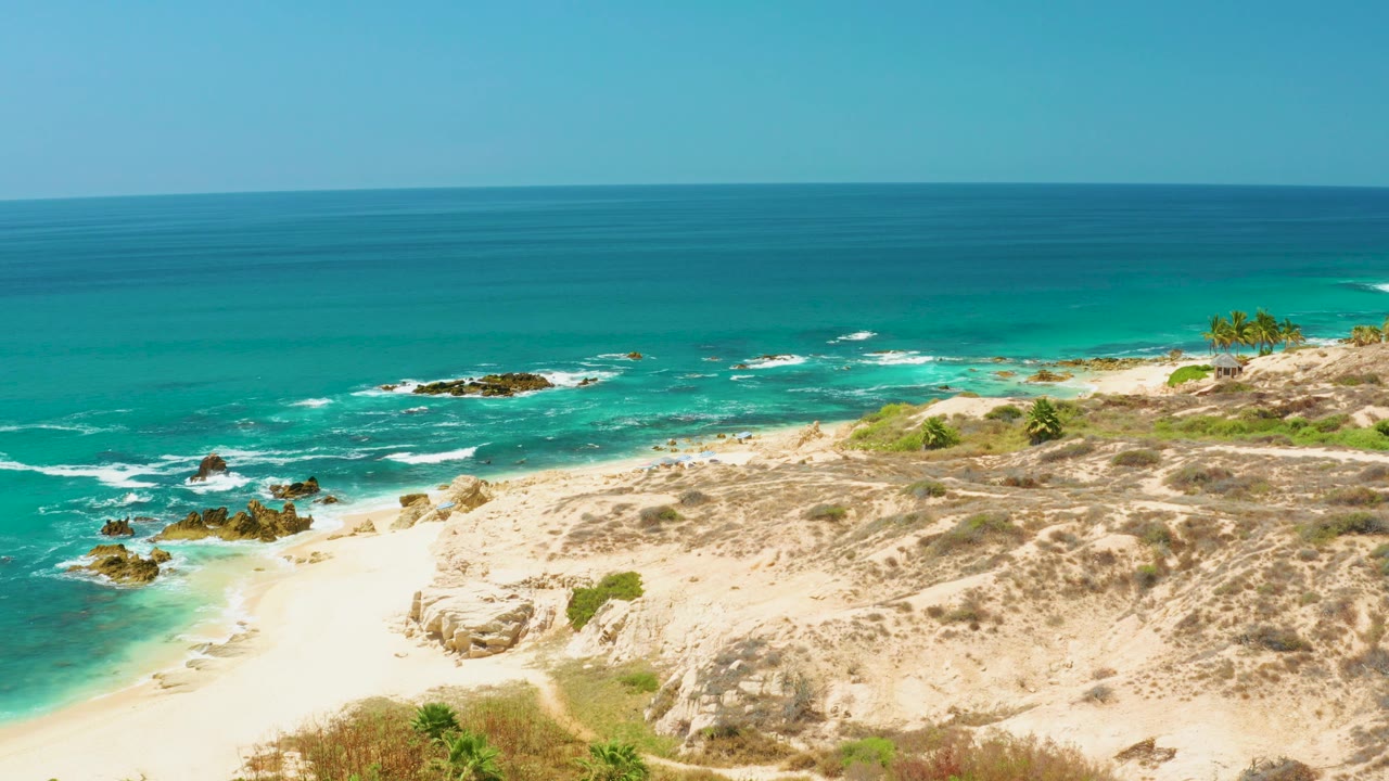 Wide elevated view of turquoise water, white surf, pale sand, rocky outcrops, dry scrub, and a clear sky at Dreams Los Cabos, with no visible people.