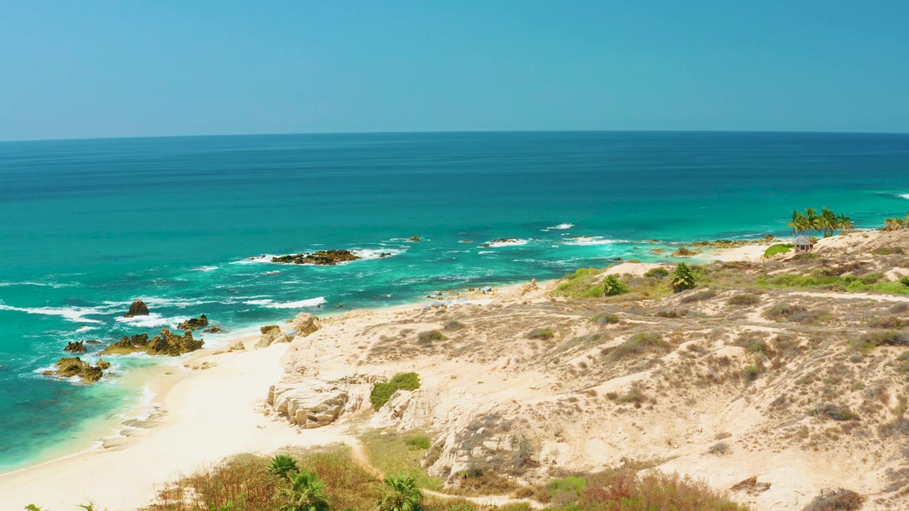 Wide coastal frame at Dreams Los Cabos with foamy water around scattered rocks, an empty sandy cove, low vegetation, and no visible people.