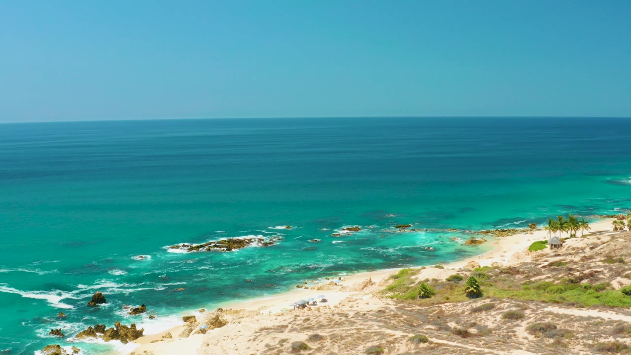 At 0:01, the Dreams Los Cabos beach remains framed from above, with whitecaps shifting near the rocks, scrub on the sandy slope, and no visible people.