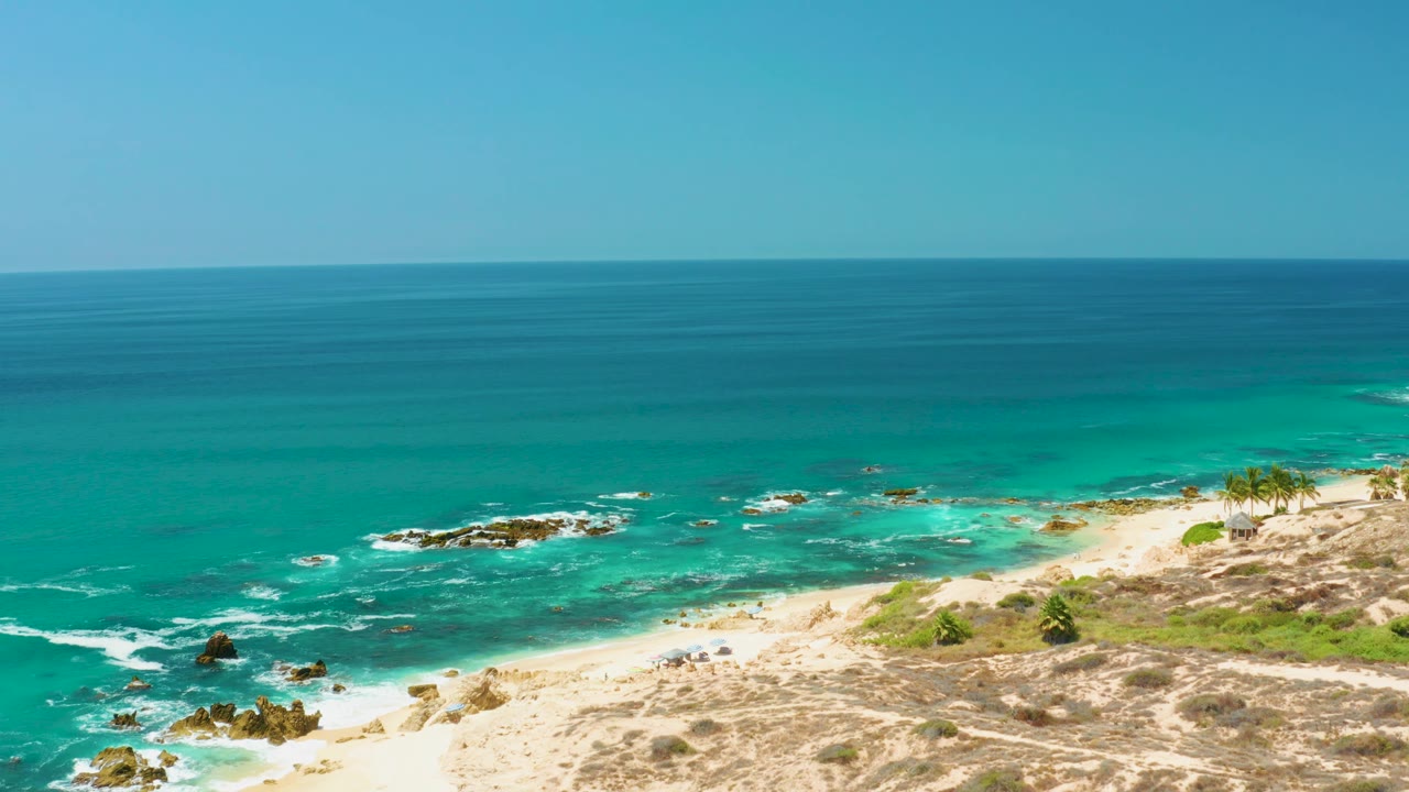 At 0:02, the elevated Dreams Los Cabos view shows a broad ocean horizon, turquoise shallows, scattered rock outcrops, sandy beach, and no visible people.