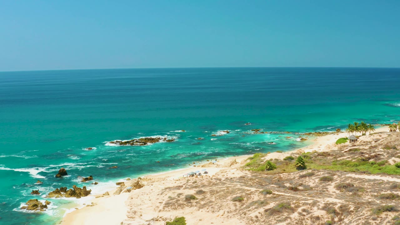 At 0:03, sandy dunes occupy more of the lower frame while turquoise waves curl toward the beach at Dreams Los Cabos, with no visible people.
