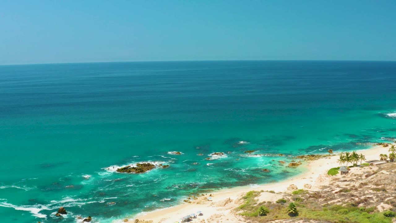Aerial view at Dreams Los Cabos showing a wide blue ocean horizon, turquoise shallows, dark rocks, sandy beach, and tiny people near the shore.