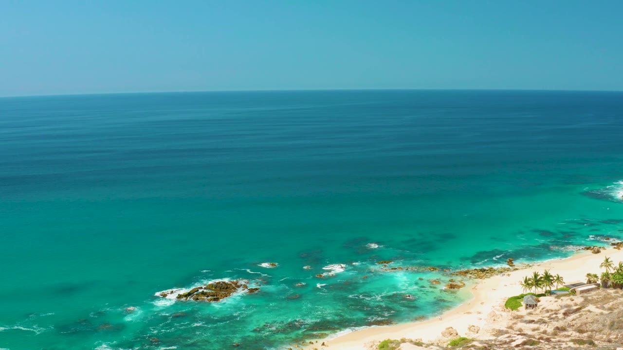 The Dreams Los Cabos shoreline remains in an elevated wide view, with breaking foam around rocks, pale sand at lower right, palm trees near the beach, and no visible people.