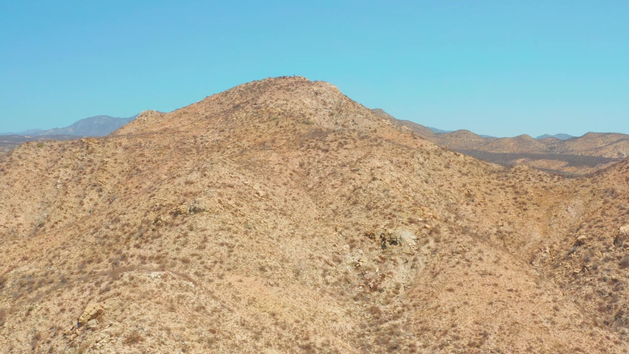 The hill remains prominent near Dreams Los Cabos, showing beige dry soil, rocky outcrops, low brush, hazy distant mountains, and no visible people.